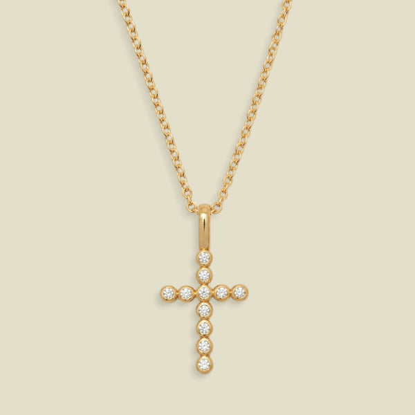 A gold cross pendant necklace adorned with small clear round bezel set gemstones, suspended from a delicate gold chain against a light beige background.