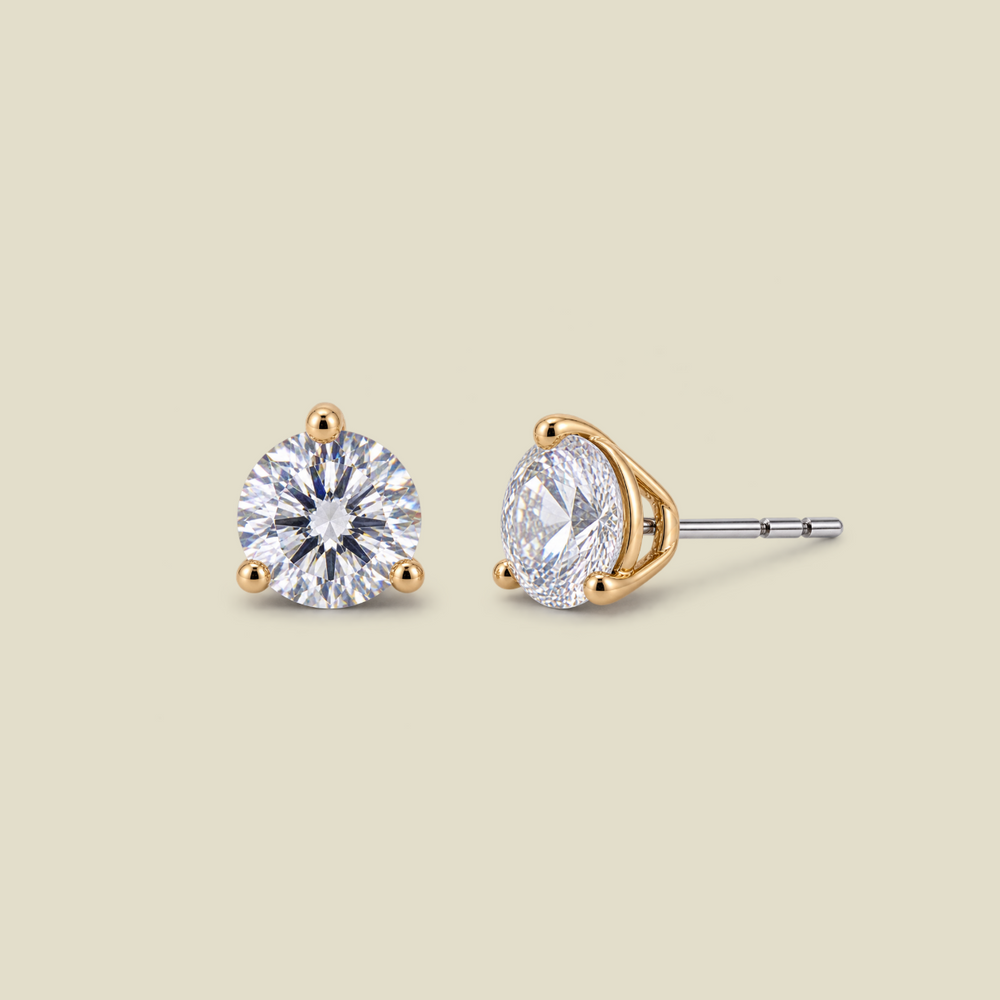 Two stud earrings on a warm beige background, one showing the front view of a round brilliant-cut clear gemstone in a gold prong setting, and the other showing the side profile with a silver-toned post and gold-toned end.