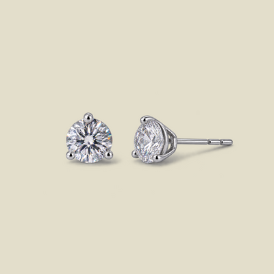 A pair of silver stud earrings with round-cut prong set clear gemstones, shown on a solid light beige background. One earring is facing forward, and the other is shown in profile.