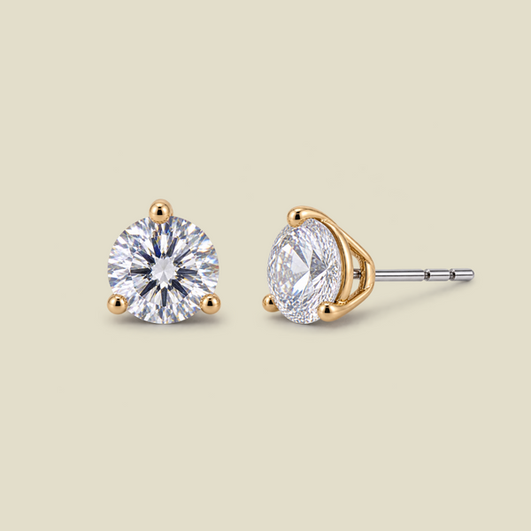 Two round-cut stud earrings in yellow gold prong settings with clear gemstones, shown on a light beige background. One earring is facing forward, and the other is angled to show the side post.