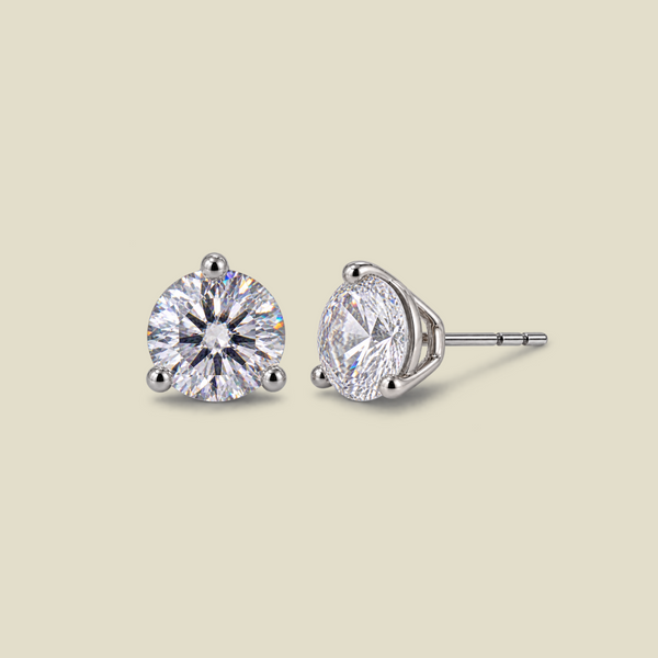 A pair of round-cut stud earrings in a silver-toned three-prong setting, shown from front and side angles on a solid light beige background.