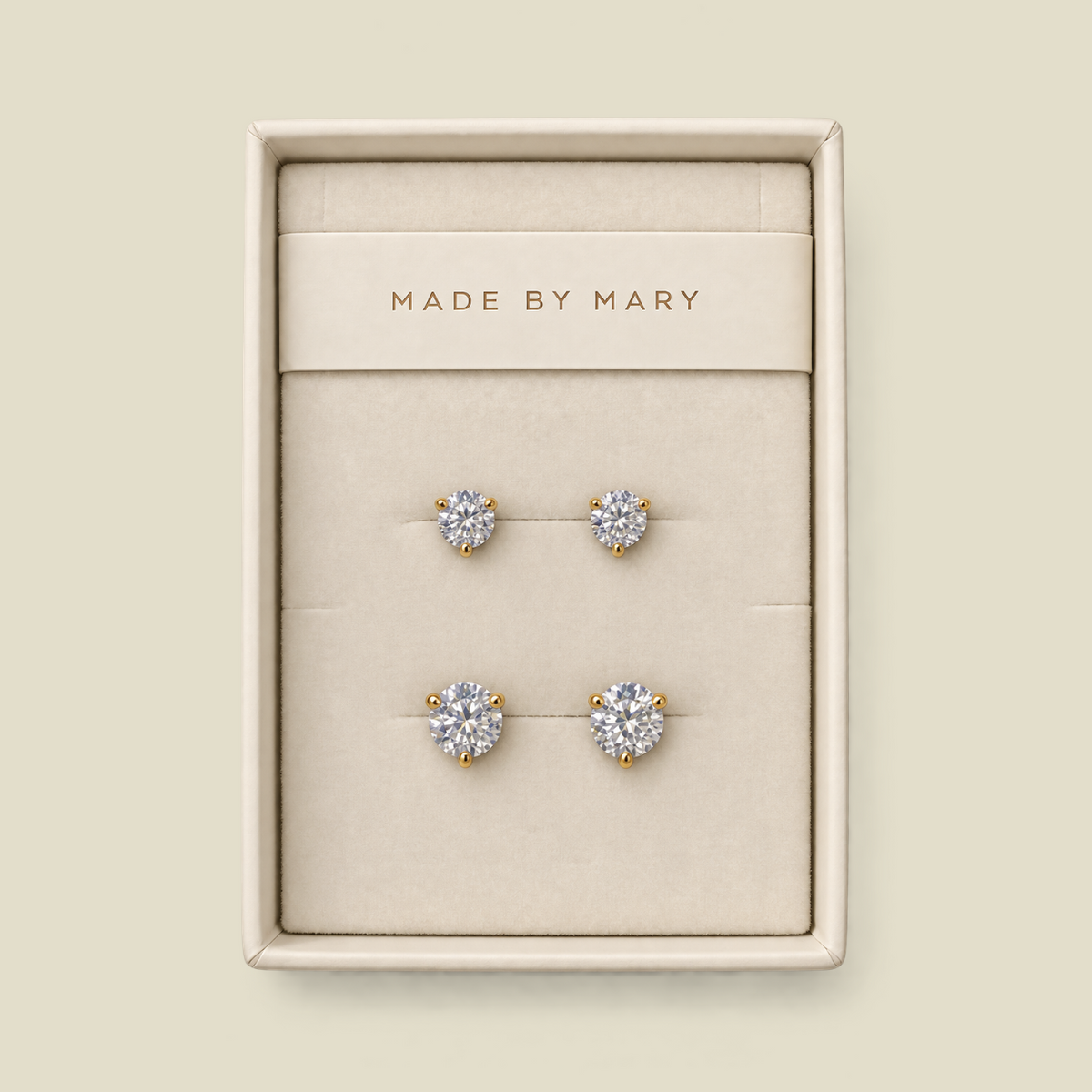 A set of four stud earrings displayed in a cream-colored jewelry box. The earrings feature round clear gemstones in yellow gold settings, with one smaller pair and one larger pair.