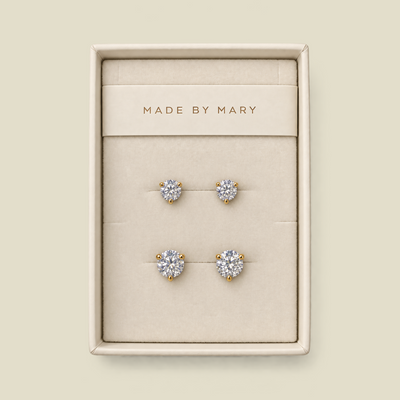 A set of four stud earrings displayed in a cream-colored jewelry box. The earrings feature round clear gemstones in yellow gold settings, with one smaller pair and one larger pair.