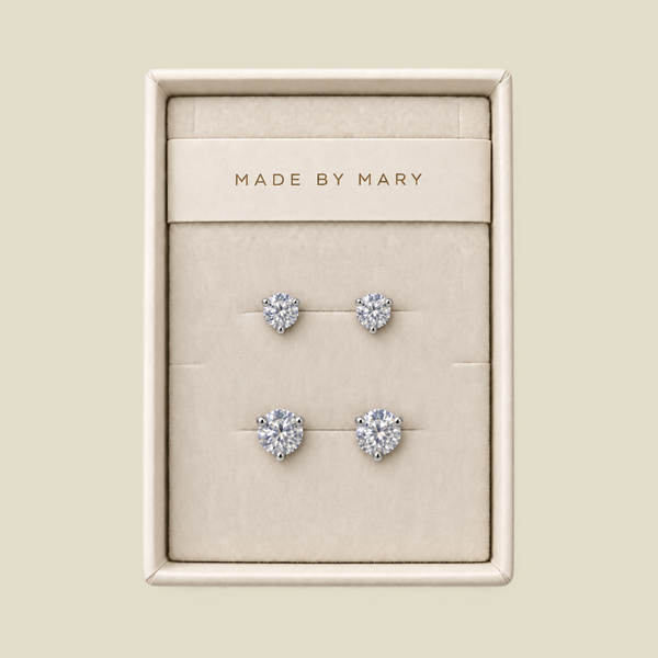A set of four stud earrings displayed in a cream-colored jewelry box. The earrings feature round clear gemstones in silver settings, with one smaller pair and one larger pair.