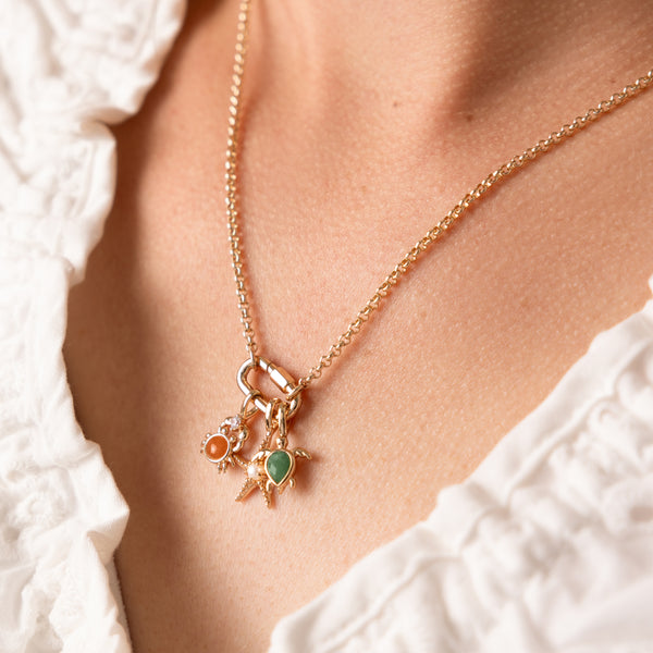 A close-up of a delicate gold chain necklace worn on a model's neck, featuring three small charms: one with a round orange stone in the shape of a crab, one in the shape of a starfish with a pearl in the center, and another shaped like a turtle with a green stone, all set in gold.