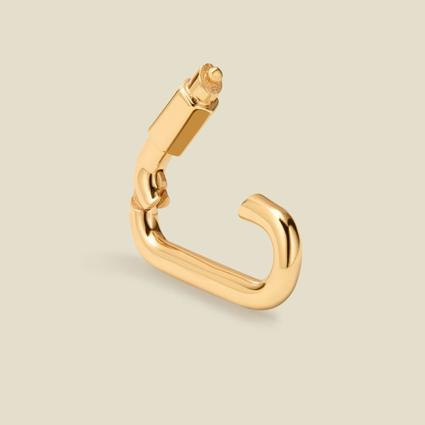 A single gold-toned carabiner shaped accessory with a polished finish, shown on a plain warm beige background.