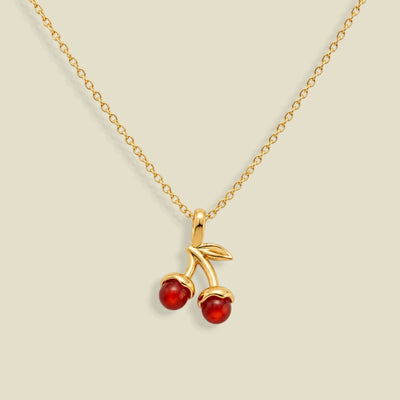 A gold chain necklace featuring a cherry pendant with two red gemstone cherries and a small gold leaf, displayed on a light beige background.