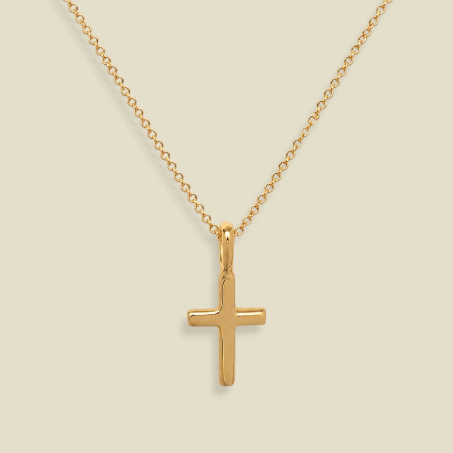 A dainty gold chain with a small gold cross charm, set against a solid soft beige background.