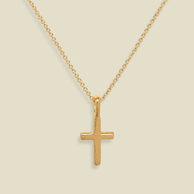A dainty gold chain with a small gold cross charm, set against a solid soft beige background.