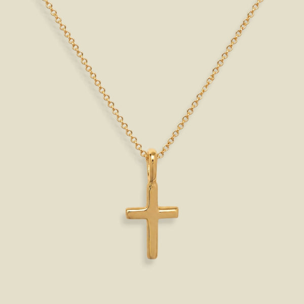 A dainty gold chain with a small gold cross charm, set against a solid soft beige background.