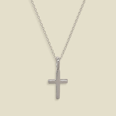 A silver cross pendant necklace on a delicate silver chain, displayed against a solid light beige background.