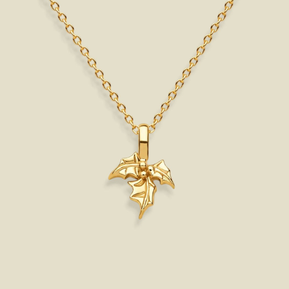 A gold holly flower pendant necklace on a delicate gold chain, presented against a solid light beige background.
