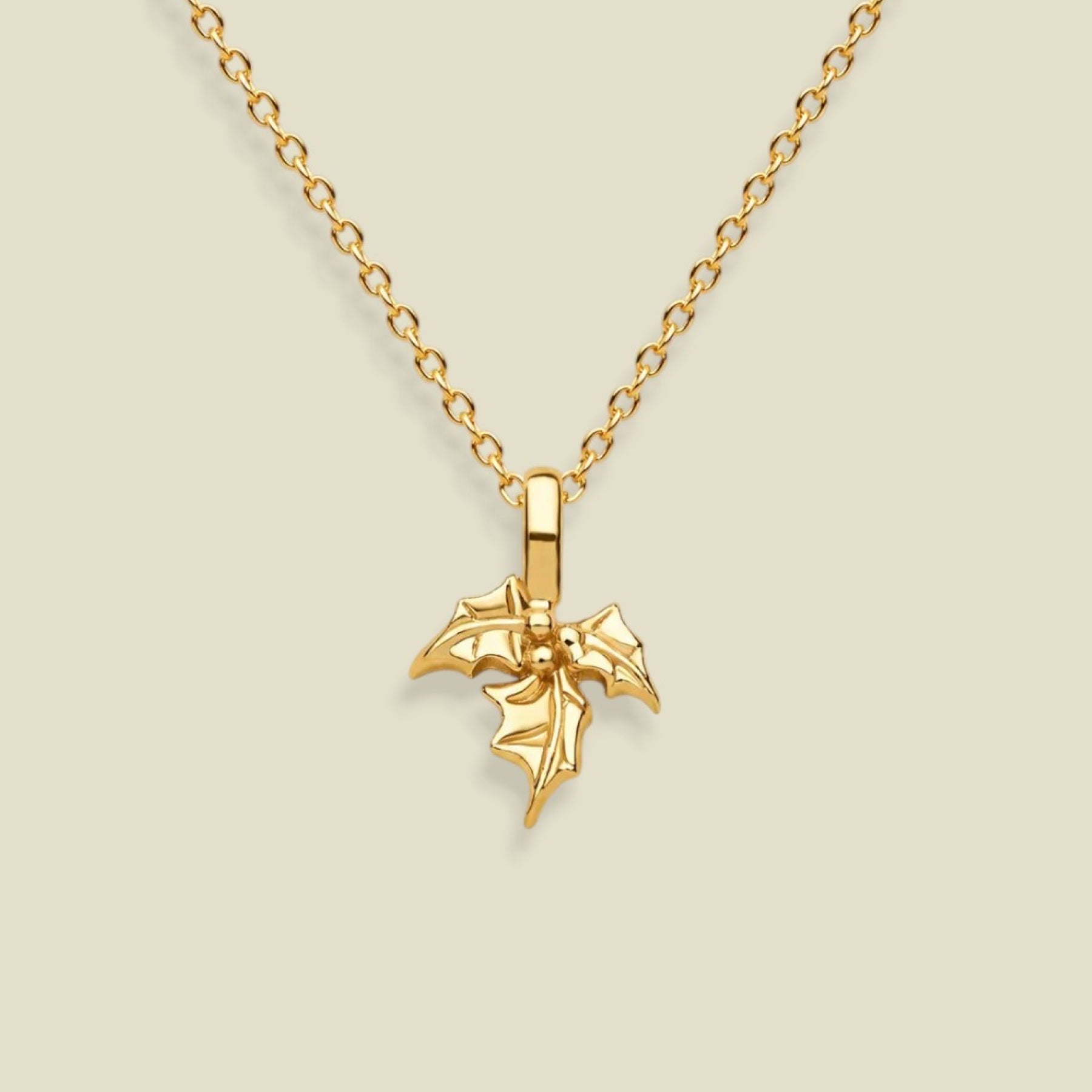 A gold holly flower pendant necklace on a delicate gold chain, presented against a solid light beige background.