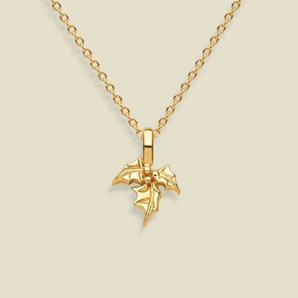 A gold holly flower pendant necklace on a delicate gold chain, presented against a solid light beige background.