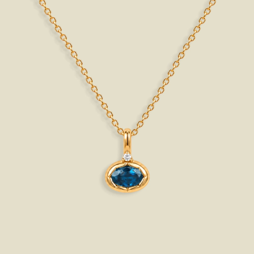 A gold chain necklace with a pendant featuring an oval cut blue gemstone in a gold bezel setting, accented by a small clear stone on the top, set against a solid light beige background.