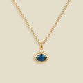 A gold chain necklace with a pendant featuring an oval cut blue gemstone in a gold bezel setting, accented by a small clear stone on the top, set against a solid light beige background.