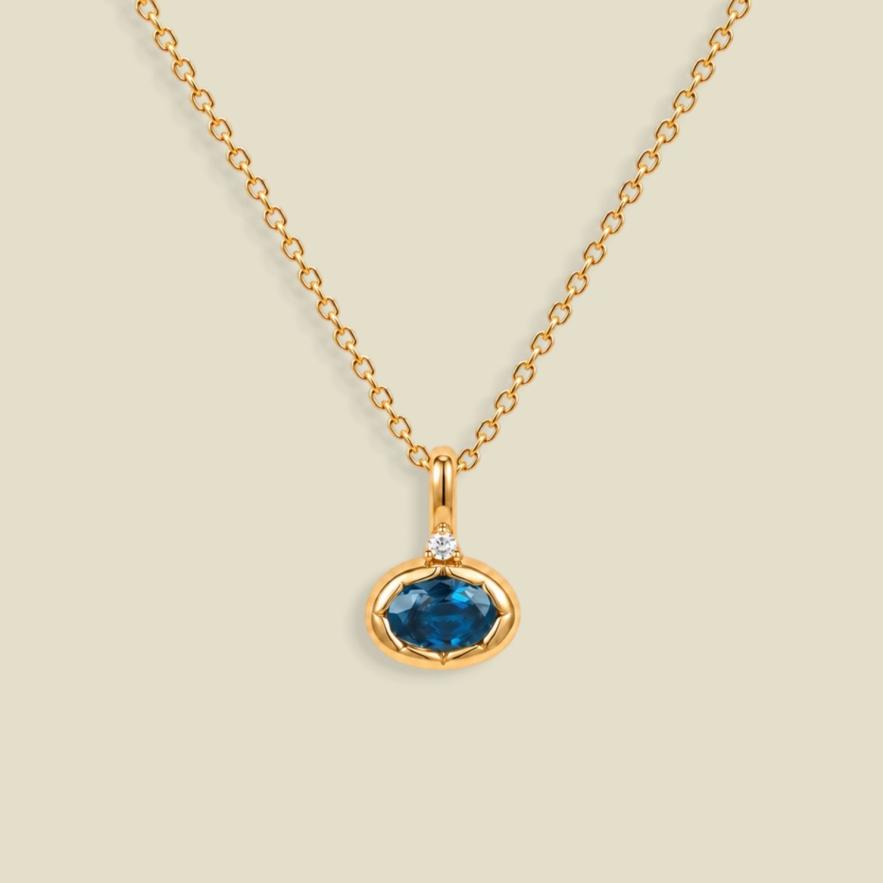 A gold chain necklace with a pendant featuring an oval cut blue gemstone in a gold bezel setting, accented by a small clear stone on the top, set against a solid light beige background.
