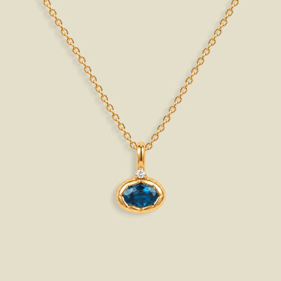 A gold chain necklace with a pendant featuring an oval cut blue gemstone in a gold bezel setting, accented by a small clear stone on the top, set against a solid light beige background.