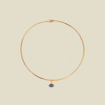 A delicate gold wire hoop necklace with a small oval blue gemstone pendant, displayed on a solid light beige background.