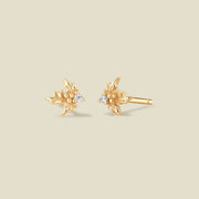 A pair of gold holly stud earrings with small clear gemstones in the center, shown on a solid light beige background.