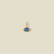 A gold oval pendant charm with a blue bezel-set oval gemstone and a clear accent stone on top, set against a solid beige background.