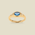 A gold signet ring with a blue center oval bezel-set gemstone, and two small clear accent stones on either side, set against a solid beige background.