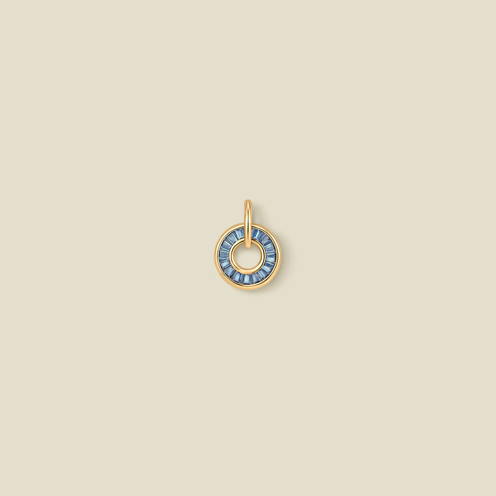 A circular gold pendant featuring blue baguette-cut stones around a hollow center, displayed against a solid light beige background.