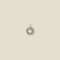 A circular gold pendant featuring blue baguette-cut stones around a hollow center, displayed against a solid light beige background.