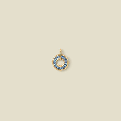 A circular gold pendant featuring blue baguette-cut stones around a hollow center, displayed against a solid light beige background.