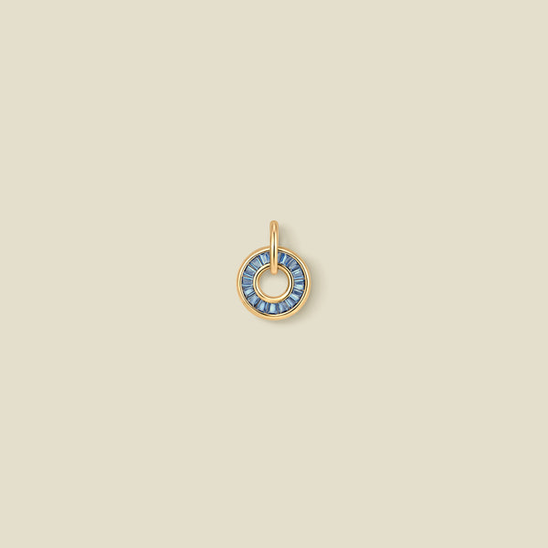 A circular gold pendant featuring blue baguette-cut stones around a hollow center, displayed against a solid light beige background.