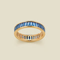 A gold eternity ring featuring a continuous channel setting of baguette-cut blue gemstones, shown on a solid light beige background.