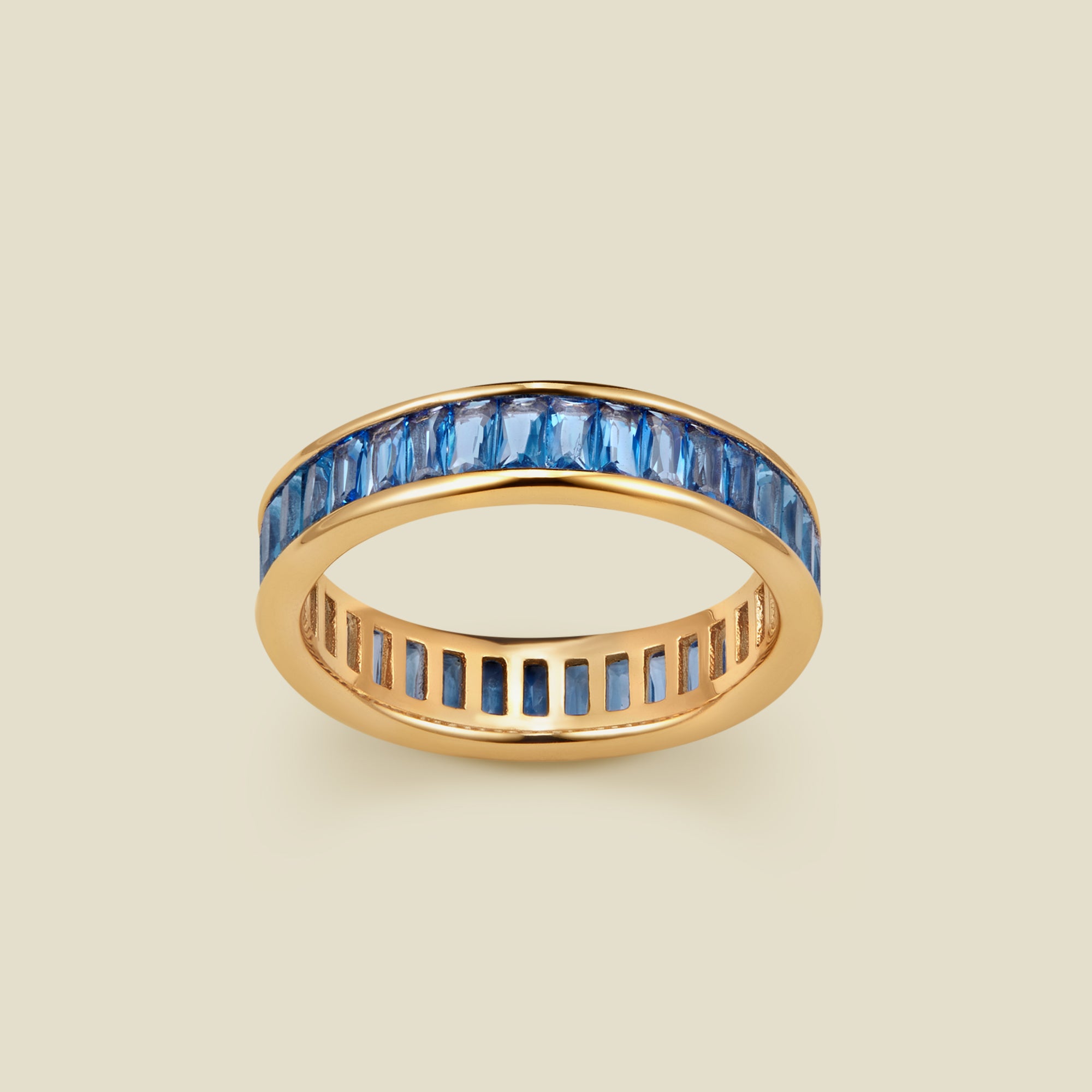 A gold eternity ring featuring a continuous channel setting of baguette-cut blue gemstones, shown on a solid light beige background.