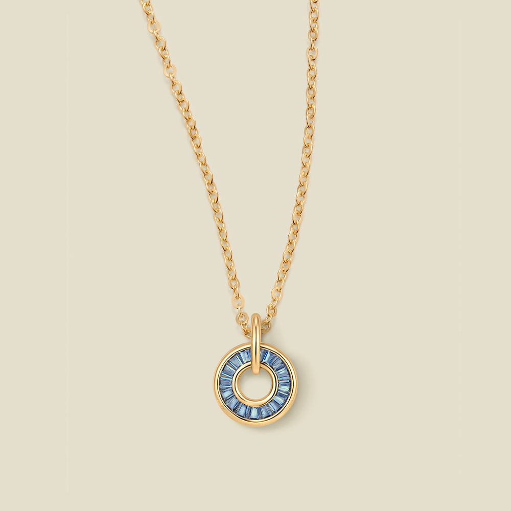 A gold chain necklace with a circular pendant featuring blue baguette-cut stones around a hollow center, displayed against a solid light beige background.