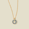 A gold chain necklace with a circular pendant featuring blue baguette-cut stones around a hollow center, displayed against a solid light beige background.