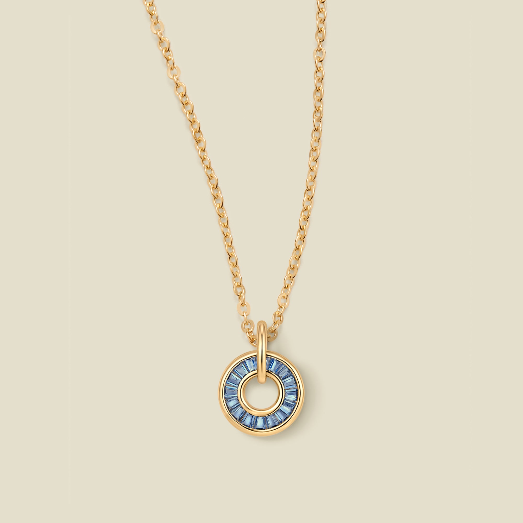A gold chain necklace with a circular pendant featuring blue baguette-cut stones around a hollow center, displayed against a solid light beige background.