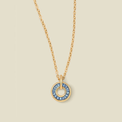 A gold chain necklace with a circular pendant featuring blue baguette-cut stones around a hollow center, displayed against a solid light beige background.