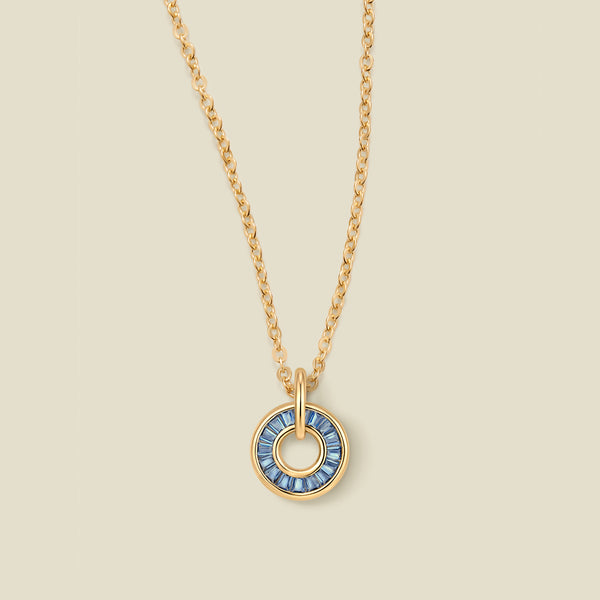A gold chain necklace with a circular pendant featuring blue baguette-cut stones around a hollow center, displayed against a solid light beige background.