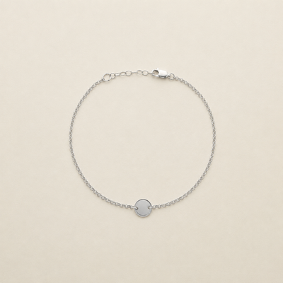 A delicate silver chain anklet with a small, polished circular disc charm in the center, displayed on a warm cream-colored background.