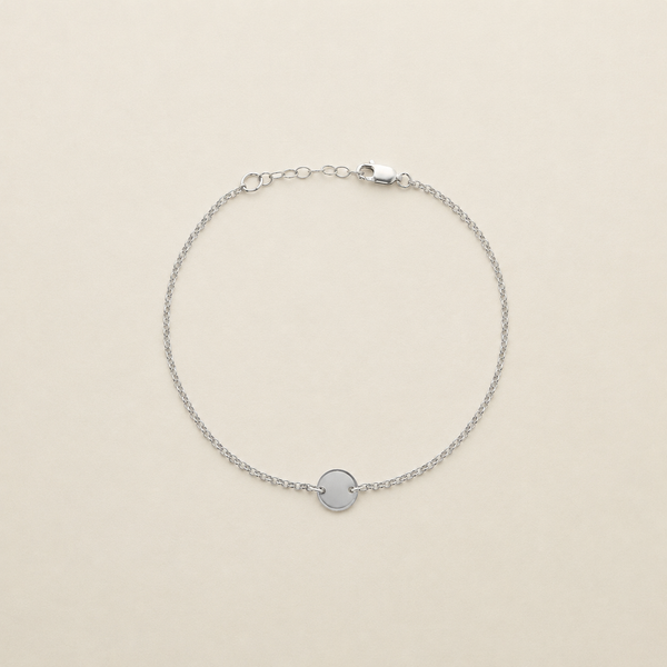 A delicate silver chain anklet with a small, polished circular disc charm in the center, displayed on a warm cream-colored background.