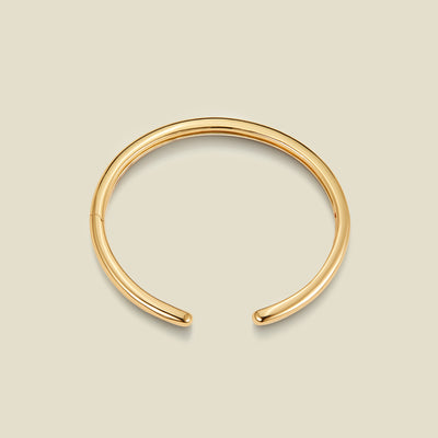 A polished gold-toned open cuff bangle bracelet, displayed on a solid light beige background.