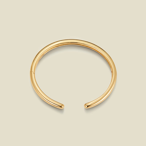 A polished gold-toned open cuff bangle bracelet, displayed on a solid light beige background.