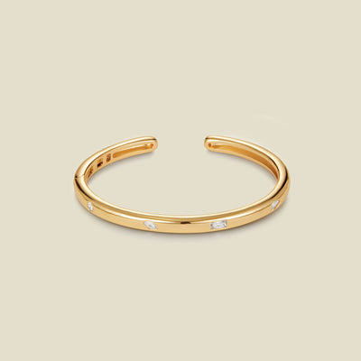 A polished gold open cuff bracelet with small clear rectangular stones set along the front, displayed on a solid light beige background.