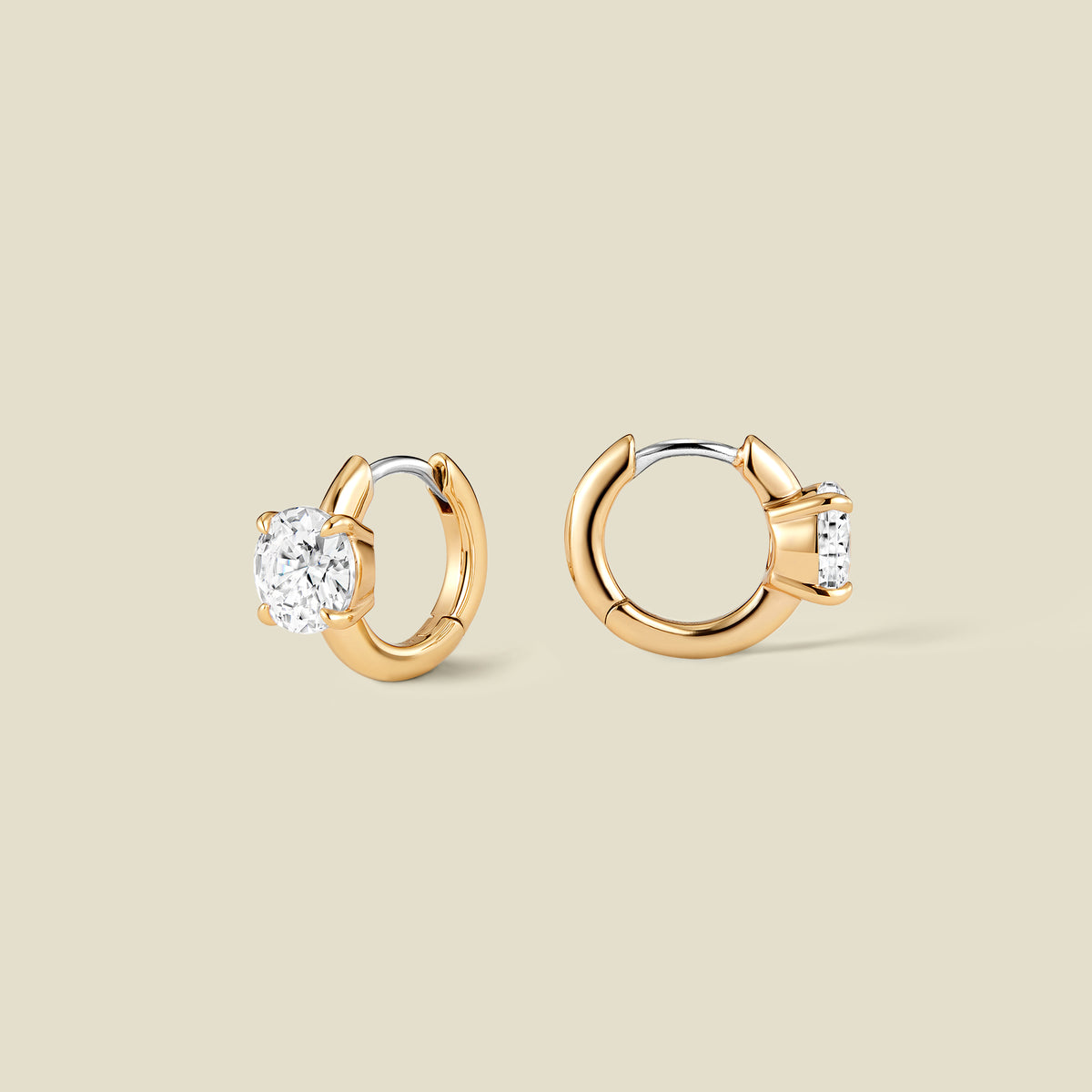 A pair of gold huggie hoop earrings with a single round-cut clear gemstone in a prong setting, shown on a solid beige background.