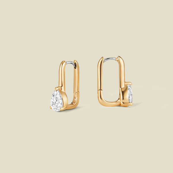 A pair of gold drop hoop earrings with pear-cut clear gemstones, shown from two angles on a solid beige background.
