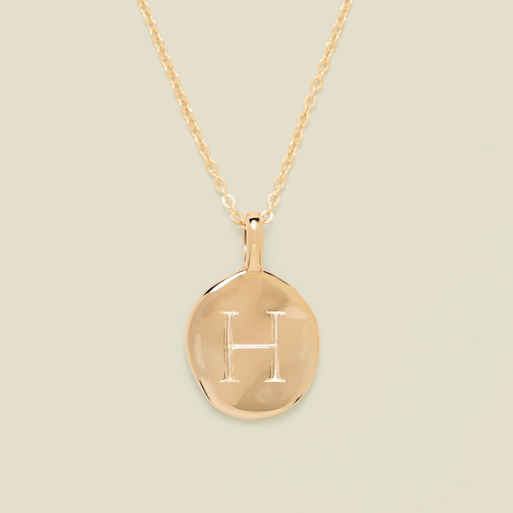 Gold necklace with a round pendant engraved with the letter 'H' on a beige background