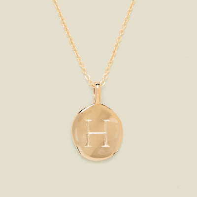 Gold necklace with a round pendant engraved with the letter 'H' on a beige background