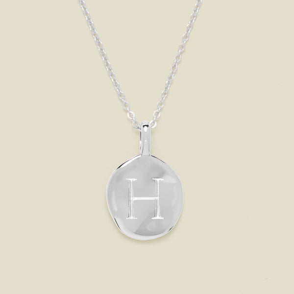 Silver necklace with a round pendant engraved with the letter 'H' on a beige background