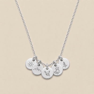 A silver chain necklace with five small round engraved disc charms, each featuring a different simple symbol (sun, mountain, butterfly, teddy bear, and a pine tree), displayed on a solid beige background.