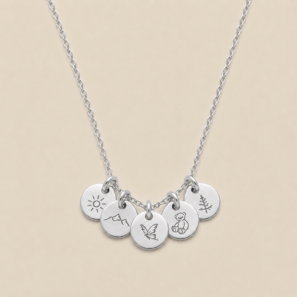 A silver chain necklace with five small round engraved disc charms, each featuring a different simple symbol (sun, mountain, butterfly, teddy bear, and a pine tree), displayed on a solid beige background.