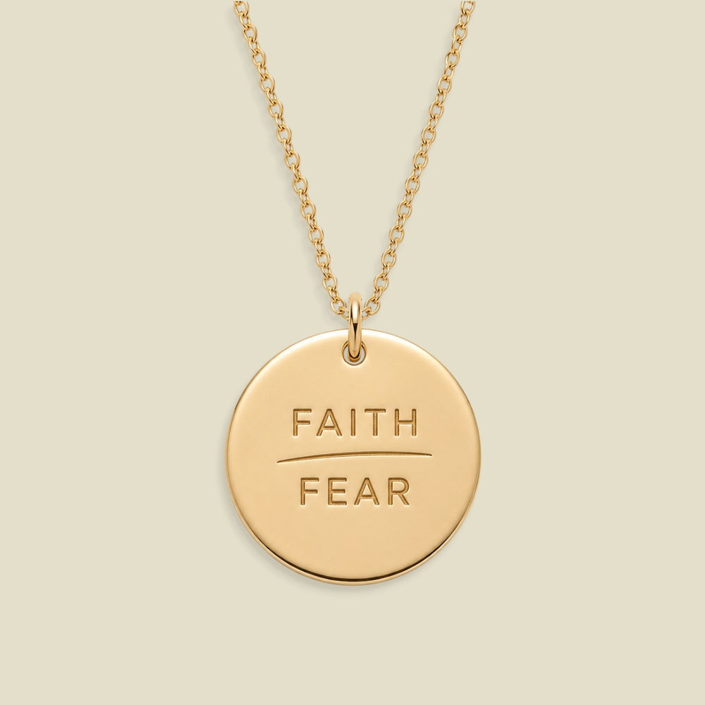 A gold disc pendant necklace engraved with the words 'FAITH' and 'FEAR' in capital letters, separated by a horizontal line, suspended from a delicate gold chain against a solid light beige background.